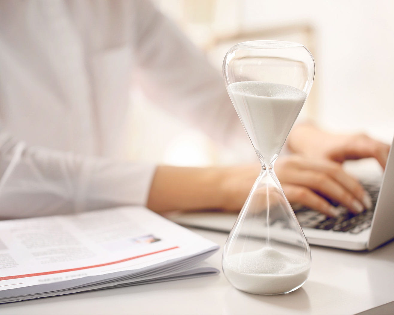 A close-up of an hourglass beside a person typing on a laptop, with papers laid out on a desk.