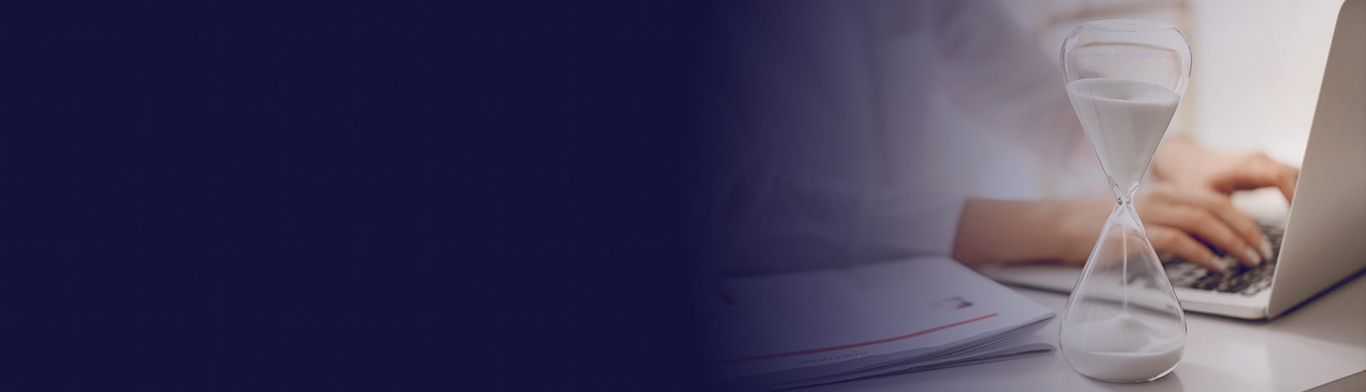 A close-up of an hourglass beside a person typing on a laptop, with papers laid out on a desk.