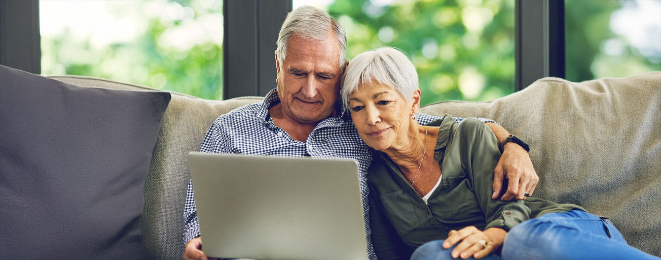 A senior couple sits closely on a couch, sharing a laptop.