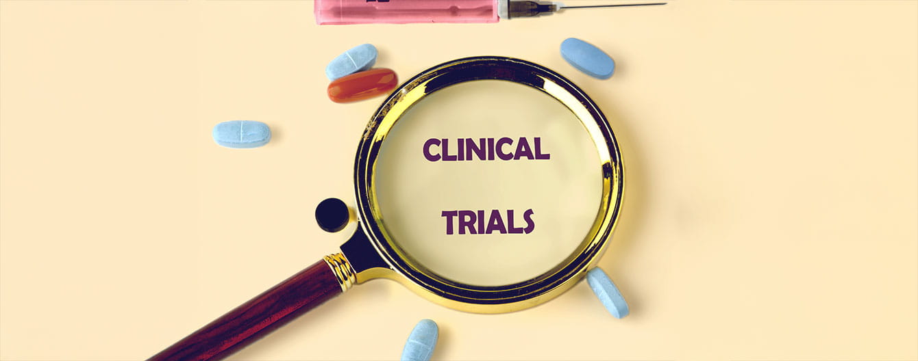A magnifying glass focuses on the words "CLINICAL TRIALS," surrounded by colorful pills and a syringe on a beige background.