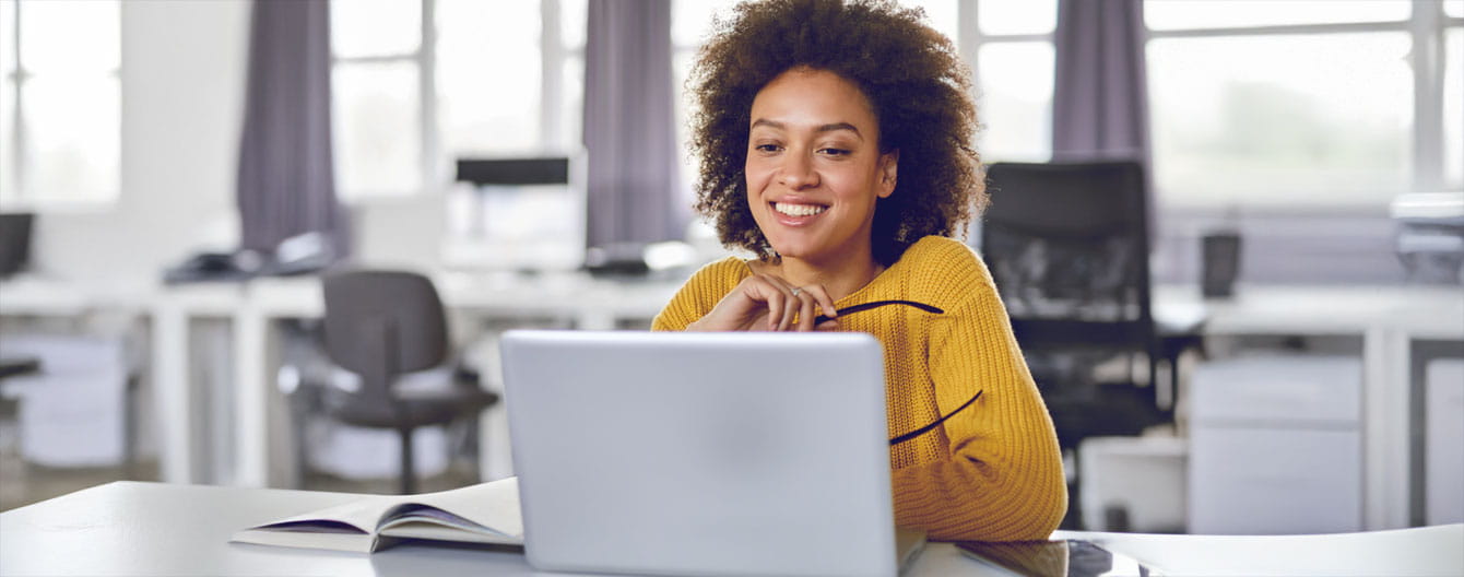 A woman in a yellow sweater sits at a desk, intently looking at her laptop.