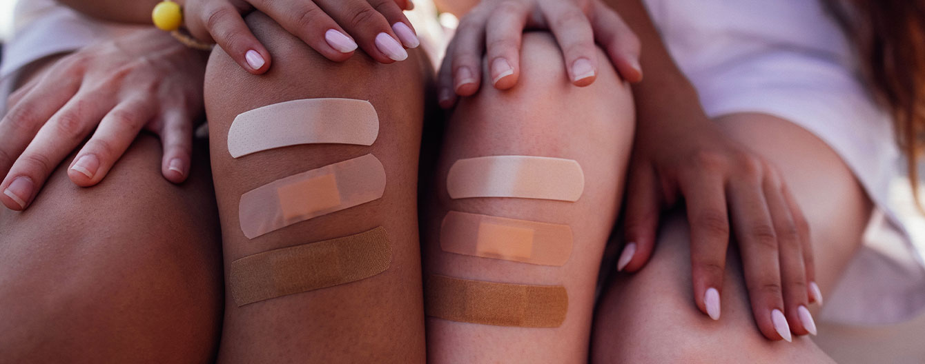 Close-up of two women’s knees with various skin-toned bandages, showcasing inclusivity in care.