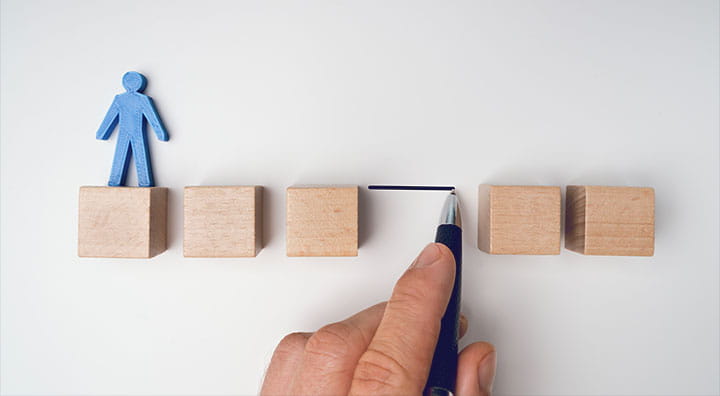 A blue figure stands on a wooden block while a hand draws a line with a pen in between the gap of the blocks.