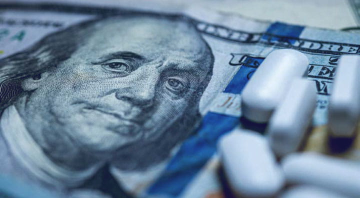 A close-up of white pills scattered on a blurred background of U.S. hundred-dollar bills.