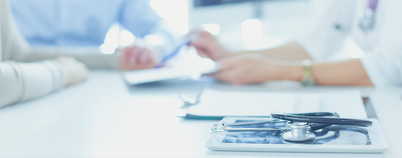 A close-up of a doctor and patient in a hospital setting, with a stethoscope and laptop on the desk.